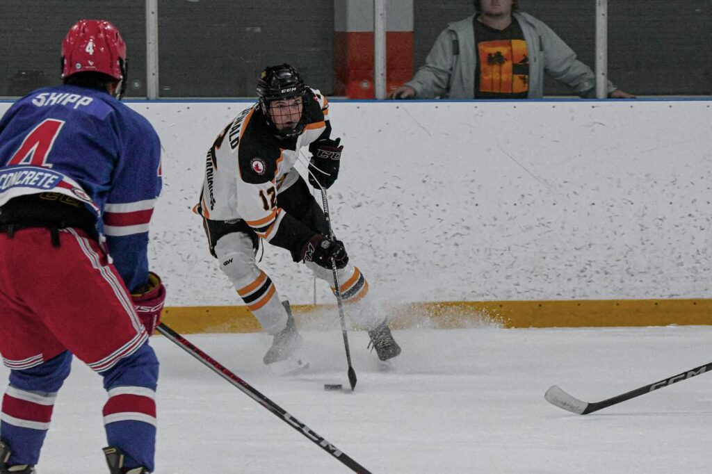 Goderich Flyers player Kaeden MacDonald, wearing a white and black jersey with the number 17, is seen controlling the puck along the boards, kicking up ice as he makes a sharp turn. A Kincardine Bulldogs player in a blue and red uniform, number 4, faces him, preparing to defend. In the background, a spectator watches the play closely from behind the glass.