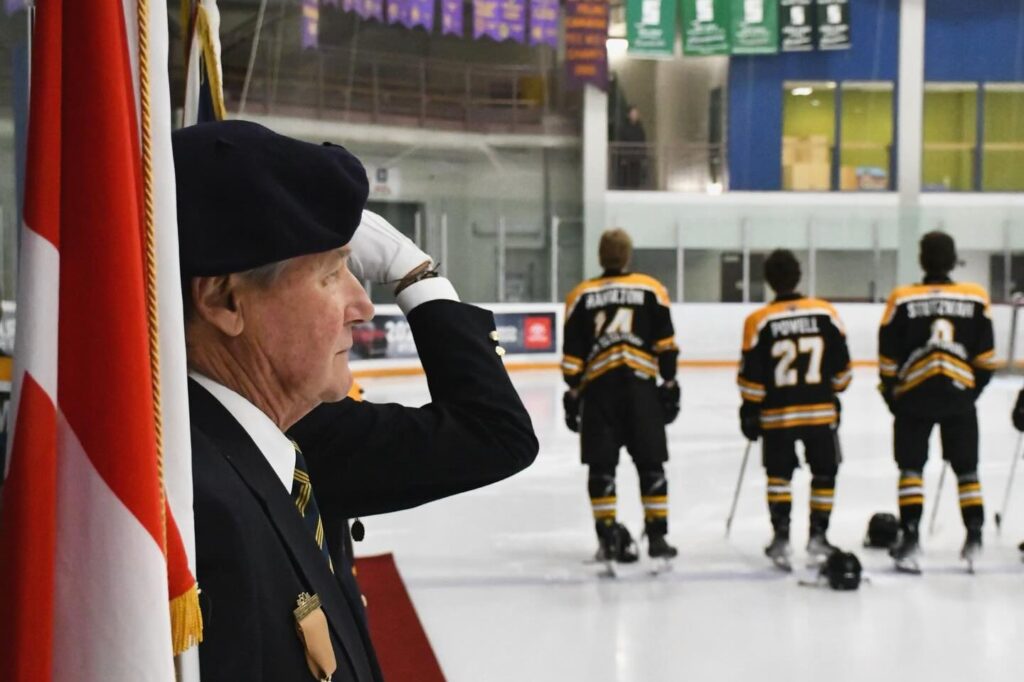 A veteran in a black beret and suit jacket stands at attention, saluting the Canadian flag during a pre-game ceremony at an ice rink. In the background, three Goderich Flyers players, seen from behind, stand respectfully on the ice with their helmets placed in front of them. The players wear black and gold jerseys, with visible names and numbers, including “Hamilton” (#14), “Powell” (#27), and “Stutzman” (#9). The arena features banners hanging from the ceiling and the Canadian flag overhead, creating a solemn and patriotic atmosphere.