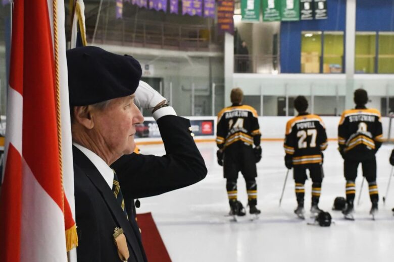 A veteran in a black beret and suit jacket stands at attention, saluting the Canadian flag during a pre-game ceremony at an ice rink. In the background, three Goderich Flyers players, seen from behind, stand respectfully on the ice with their helmets placed in front of them. The players wear black and gold jerseys, with visible names and numbers, including “Hamilton” (#14), “Powell” (#27), and “Stutzman” (#9). The arena features banners hanging from the ceiling and the Canadian flag overhead, creating a solemn and patriotic atmosphere.