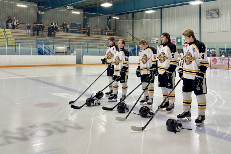 A group of Goderich Flyers players, dressed in white jerseys with gold and black trim, stand in a line on the ice with their helmets placed in front of them. They hold their hockey sticks vertically, resting on the ice, as they face the stands during a moment of respect or anthem. The arena is sparsely populated, with a few spectators seated in the bleachers behind them. The players are focused and still, creating a solemn, unified image on the rink.